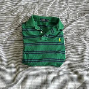 Grön randig pikétröja från Ralph Lauren - Säljer en snygg grön pikétröja från Ralph Lauren med mörkblå ränder och det klassiska gula broderade logotypen på bröstet. Perfekt för en stilren och avslappnad look. Tröjan har korta ärmar och en klassisk krage.