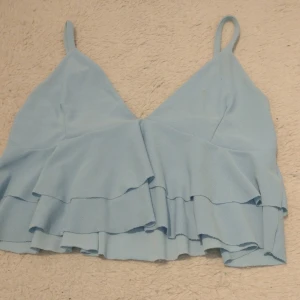 Ljusblå volang linnetopp - Säljer en ljusblå croppad linnetopp från PrettyLittleThings med tunna axelband och dubbla volanger framtill. Står ingen storlek men passar mig som har XS/S💗 Har sytt banden kortare, men går att sprätta upp om man vill ha banden längre! (sista bild)