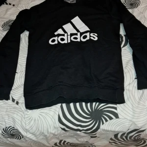 Svart sweatshirt från Adidas - Svart sweatshirt från Adidas med klassisk vit logga framtill. Tröjan har rund halsringning och långa ärmar. Perfekt för en sportig och avslappnad stil.