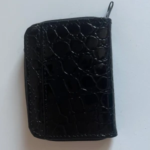 Svart plånbok med krokodilpräglat mönster - Säljer en svart plånbok med glansig krokodilpräglad yta. Plånboken har dragkedja runtom och flera fack inuti för kort och sedlar. Perfekt storlek för att ha med sig i väskan eller fickan.