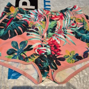 Blommiga shorts från Hampton Republic - Säljer ett par färgglada shorts från Hampton Republic med tropiskt blommönster i grönt, blått, rött och gult på en rosa bas. Shortsen har dragsko i midjan och mjukt bomullsmaterial. Perfekta för sommardagar! Nya aldrig använda.  Storlek 146-152 