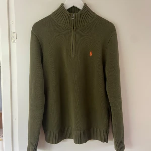 Polo Ralph lauren half zip - Säljer en olivgrön stickad tröja från Polo Ralph Lauren med halv dragkedja och hög krage. Tröjan har lång ärm och den klassiska broderade loggan i orange på bröstet. Perfekt för lager-på-lager under kyliga dagar.
