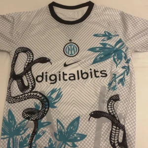 Inter Milan bortatröja med ormmönster - Säljer en vit Inter Milan fotbollströja från Nike med svart ormmönster och blåa blad. Tröjan har svart krage och digitalbits som sponsortryck på bröstet. Perfekt för dig som vill sticka ut på planen eller läktaren.