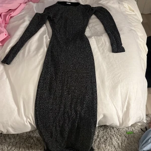 Svart långklänning med glitter från Gina tricot  - Säljer en svart långklänning med vita prickar och långa ärmar. Klänningen har en snygg v-ringning och rynkade detaljer framtill. Perfekt för dig som gillar en figurnära passform och vill sticka ut lite extra.