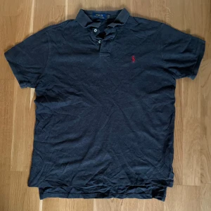 Ralph Lauren Piké  - Säljer en klassisk mörkgrå pikétröja från Polo Ralph Lauren med röd broderad logga på bröstet. Tröjan har korta ärmar, krage och knappar vid halsen. Perfekt för en stilren och avslappnad look.