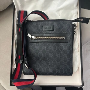 Gucci väska - Svart äkta Gucci axelremsväska med justerbar rem i rött och blått. Väskan har en dragkedja upptill och en mindre dragkedjeförsedd ficka på framsidan. Levereras med originalkartong och dustbag.  Pris kan förhandlas vid snabb affär
