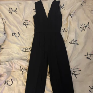 Svart byxdress med djup v-ringning - Säljer en svart byxdress med djup v-ringning och breda axelband. Byxdressen har vida ben och markerad midja för en snygg siluett. Perfekt för dig som vill ha en stilren och trendig look.