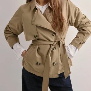 Beige trenchcoat med bälte och knappar - Snygg beige trenchcoat med dubbelknäppning och bälte i midjan. Jackan har klassisk krage, långa ärmar och knappar framtill. 
