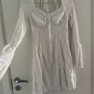Vit spetsklänning med trumpetärmar - Superfin vit kortklänning med spetsdetaljer och trumpetärmar. Klänningen har markerad byst med knyt framtill och v-ringad hals. Perfekt för sommarens tillfällen eller student. Aldrig använd.