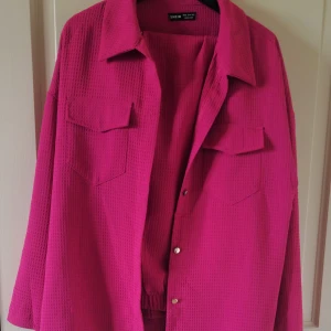 Rosa overshirt med byxor från SHEIN - Säljer en starkt rosa overshirt med byxor från SHEIN i storlek XL. Jackan har två bröstfickor med lock, klassisk krage och knäppning framtill. Materialet är strukturerat och ger en trendig look. Perfekt att slänga över en t-shirt för en färgklick i vardagen.