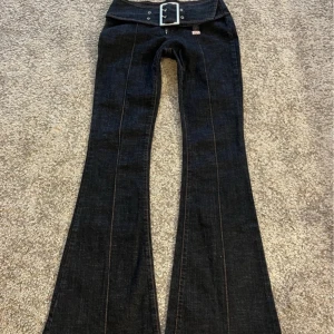 Mörkblå bootcut jeans med bälte från Miss Sixty - Säljer ett par mörkblå bootcut jeans från Miss Sixty med bred låg midja och stort bälte med spänne. Jeansen har markerade sömmar och en snygg utsvängd passform. Perfekta för dig som gillar retrostil och vill sticka ut.