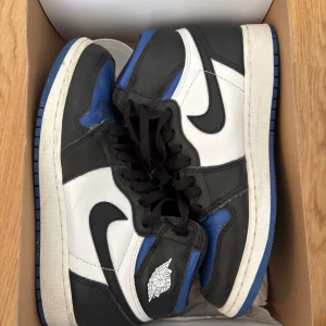 Nike Air Jordan 1 Retro High OG GS svart/vit/blå - Säljer ett par Nike Air Jordan 1 Retro High OG GS i svart, vitt och blått. Klassisk high-top modell med svart swoosh, blå detaljer på tå och häl samt vit panel. Skorna har snörning och den ikoniska Air Jordan-loggan på sidan. No crease skydd följer även med. 