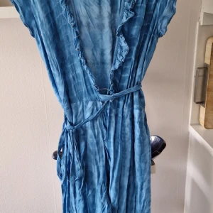 Blå omlottklänning med volang - Säljer en blå omlottklänning med knytband i midjan och volangdetaljer längs den v-ringade urringningen. Klänningen har korta ärmar och ett mjukt, lätt material som ger ett avslappnat intryck.