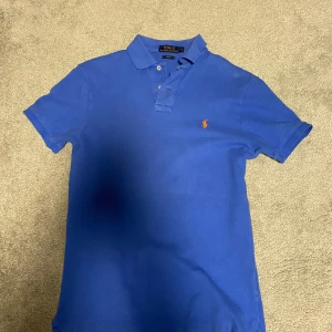 Blå pikétröja från Polo Ralph Lauren - Säljer en klassisk blå pikétröja från Polo Ralph Lauren i slim fit-modell. Tröjan har korta ärmar, krage med knappar och den ikoniska broderade loggan i orange på bröstet. Perfekt för en stilren och sportig look.
