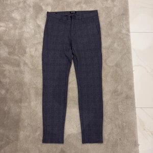 Grå-rutiga kostymbyxor från Only & Sons - Snygga grå kostymbyxor från Only & Sons med diskret rutmönster. Byxorna har normal passform, bälteshällor och knäppning fram. Perfekta för en stilren look och passar till skjorta eller t-shirt.