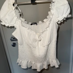 Vit volangtopp med smock och knyt - Supersöt vit topp från Gina Tricot med smockad midja, volangdetaljer vid axlar och nederkant samt knyt framtill. Toppen har korta puffärmar och är tillverkad i ett strukturerat, stretchigt material. Använd 1 gång. 💕