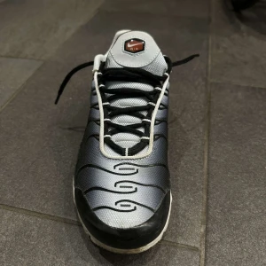 Nike tn sneakers i svart och grå - Säljer ett par tn sneakers i svart och grå med vita detaljer och röd swoosh. Skorna har snörning, platt sula och den klassiska vågiga designen på ovandelen. Perfekta för dig som gillar streetwear och sportig stil.