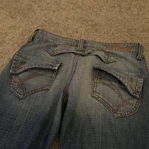 Blåa straight/loose jeansbyxor från Phard - Säljer dessa mörkblåa jeansen i loose/straight/baggy passform beroende på hur måtten passar dig. Byxorna har dekorativa bakfickor med orange sömmar och bälteshällor. Innerbenslängd: 74cm, Midjemått tvärsöver: 37cm