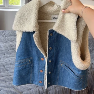Jeansväst med teddyfoder från Wrangler - Säljer en blå jeansväst från Wrangler med mjukt vitt teddyfoder och stora slag. Västen har knappar framtill och två fickor. Perfekt för lager-på-lager och ger en cool retrolook.