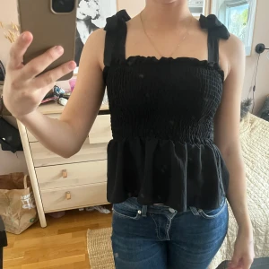 Svart smockad linnetopp med volang - Supersöt svart linnetopp med smockad överdel och volang nertill. Breda axelband med rosett-detaljer upptill. Perfekt till jeans eller kjol för en trendig look.