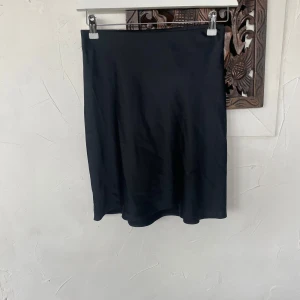 Svart satinkjol från BikBok - En stilren svart kjol i satin från BikBok. Kjolen har en rak passform och slutar ovanför knäna. Perfekt att matcha med en enkel topp eller stickad tröja. Snygg och enkel design som passar till många olika looks.