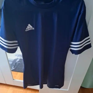 Blå Adidas fotbollströja med vita ränder - Snygg blå fotbollströja från Adidas med klassiska vita ränder på ärmarna och Adidas-logga på bröstet. Tillverkad i ett lätt och ventilerande material, perfekt för träning eller match.