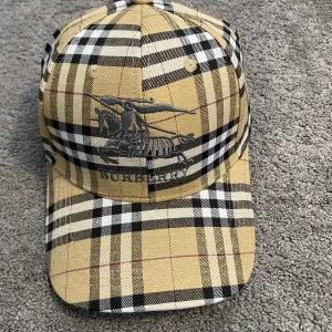 rutig keps i beige - Säljer en klassisk rutig keps i beige, svart och vitt. Kepsen har broderad logga framtill och justerbart guldspänne bak. Tillverkad i bomull med ikoniskt rutmönster och märkets detaljer invändigt.