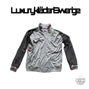 Nike vintage zip up - Snygg grå track tröja från Nike med svarta ärmar och detaljer i rött och vitt på krage och muddar. Jackan har dragkedja framtill, ribbade muddar och klassisk Nike-logga på bröstet. Perfekt för en sportig och avslappnad stil.