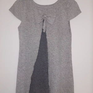 Grå stickad klänning med rosettdetalj - Grå stickad klänning med korta ärmar och en söt rosett i ryggen. Klänningen har en rak passform och är perfekt för lager-på-lager-stil. Mjuk och bekväm, passar till många tillfällen.