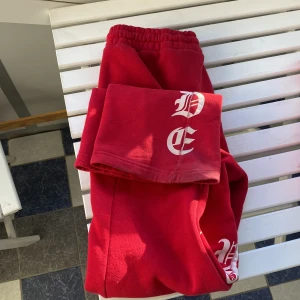 Röda mjukisbyxor med vit text  - Säljer ett par röda mjukisbyxor med resår i midjan och vit tryckt text längs ena benet. Byxorna har en avslappnad passform och är perfekta för chill dagar. Snygg streetstil och riktigt sköna att ha på sig. 