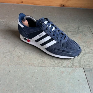 Adidas LA Trainer sneakers i marinblått - Säljer ett par klassiska Adidas LA Trainer sneakers i marinblått med vita ränder och detaljer. Skorna har snörning, mesh- och mockadetaljer samt en sula med gummidubbar för extra grepp. Perfekta för dig som gillar retrostil och sportig look. Skick 9/10 andvönda 10 gånger skriv vid frågor pris kan diskuteras vid snabb affär