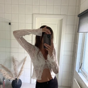 Vit spetsblus med knyt framtill - Säljer en vit, långärmad blus i spets med v-ringning och knyt framtill. Blusen är croppad och har fina volangdetaljer vid ärmslut och nederkant. Perfekt för dig som vill ha en romantisk och trendig look.