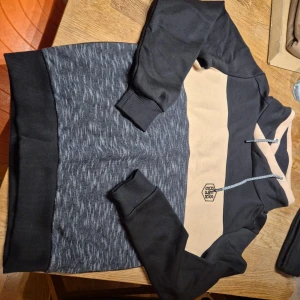 Svart och beige hoodie - Säljer en snygg hoodie i svart och beige med gråmelerad kropp. Tröjan har långa ärmar, huva med dragsko och breda muddar. Perfekt för en avslappnad stil och passar till många tillfällen.