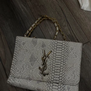 Vit axelväska med ormmönster från Saint Laurent - Säljer en snygg vit axelväska från Saint Laurent med ormmönster och guldfärgad kedja. Väskan har en stor YSL-logga framtill och är perfekt för att lyfta vilken outfit som helst.