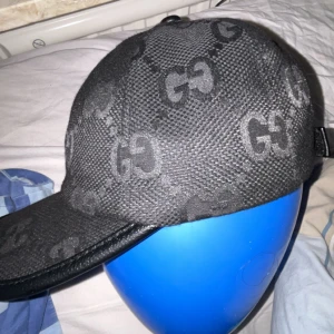 Svart keps  med GG-mönster - Säljer en svart keps  med klassiskt GG-mönster över hela kepsen. Kepsen har böjd skärm och justerbar rem baktill. Tillverkad i ett strukturerat tyg med detaljer i svart läder på skärmen.