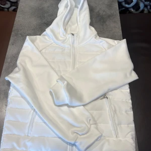 vit stilren cardigan - Säljer en stilren vit cardigan med huva och dragkedja framtill. Jackan har quiltade partier och två fickor med dragkedja. Perfekt för dig som gillar en enkel och clean look.