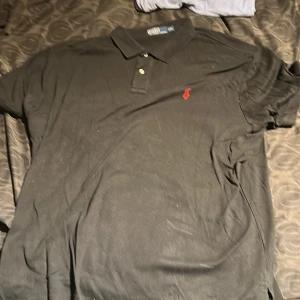 Svart pikétröja från Ralph Lauren - Klassisk svart pikétröja från Ralph Lauren med röd broderad logga på bröstet. Tröjan har korta ärmar, krage och knappar framtill. Perfekt för en stilren och avslappnad look.