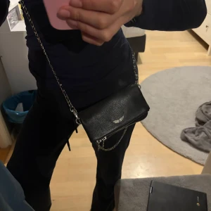 Svart liten Zadig väska - Säljer en svart axelväska i skinnimitation med silverfärgad kedjerem och dragkedja. Väskan har en liten metallogo framtill och en praktisk storlek för det viktigaste. Perfekt för dig som vill ha en stilren accessoar. Kedjan är lite rostig men inget man tänker på
