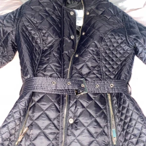 Svart quiltad jacka med bälte - Snygg svart quiltad jacka med bälte i midjan och dragkedjor i guld. Jackan har hög krage, långa ärmar och stängs med både dragkedja och knappar. Perfekt för kyligare dagar.