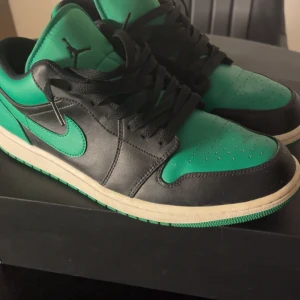 Nike Air Jordan 1 Low lucky green  - Säljer ett par Nike Air Jordan 1 Low i svart och grön med klassisk swoosh på sidan. Skorna har svart snörning, rund tå och platt sula. Perfekta för dig som gillar streetwear och vill sticka ut med färg.