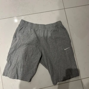 Grå träningsshorts från Nike - Gråa shorts från Nike med elastisk midja och diskret Nike-logga på ena benet. Perfekta för träning eller avslappnade dagar. Tillverkade i mjukt material för extra komfort.