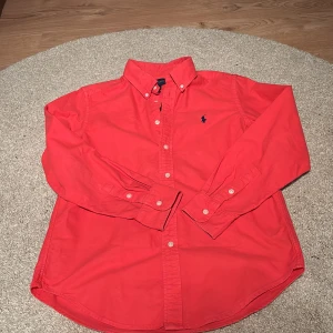 Röd skjorta från Ralph Lauren - Klassisk röd skjorta från Ralph Lauren med button-down krage och broderad logga på bröstet. Skjortan har långa ärmar och vita knappar framtill samt vid ärmsluten. Perfekt för en stilren och avslappnad look.