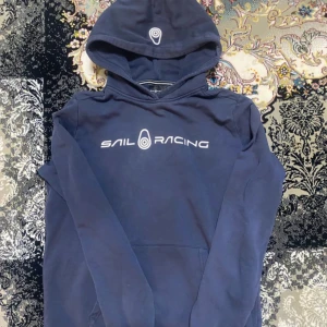 Mörkblå hoodie från Sail Racing - Säljer en mörkblå hoodie från Sail Racing med vit logga och text på bröstet. Tröjan har huva och en klassisk känguruficka framtill. Perfekt för dig som gillar en sportig och stilren look.