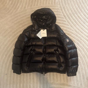 Svart dunjacka från Moncler med huva - Säljer en svart dunjacka från Moncler med huva och dragkedja. Jackan har puffig design, två sidofickor och Moncler-logga på ärmen. Insidan har ett serietidningsinspirerat foder. Perfekt för kalla dagar.
