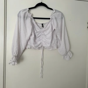 Vit croppad blus med knyt - Säljer en söt vit blus med croppad passform och långa ärmar. Blusen har volangdetaljer vid halsringningen och ärmsluten samt knyt framtill. Perfekt för vår och sommar!