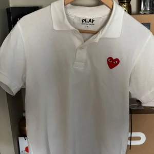 Vit piké från Comme des Garçons Play - Vit pikétröja från Comme des Garçons Play med klassisk krage och knappar. På bröstet finns det ikoniska röda hjärtat med ögon, broderat. Perfekt för en stilren och trendig look. Storlek S men sitter mer som XS