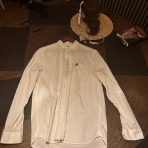 Vit skjorta från Lyle & Scott - Klassisk vit skjorta från Lyle & Scott med lång ärm och knappar framtill. Skjortan har en bröstficka med den ikoniska gula loggan broderad ovanpå. Perfekt för en clean och stilren look.