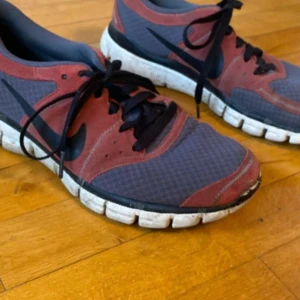 Nike sneakers i rött och grått - Säljer ett par Nike sneakers i rött och grått med svart swoosh och svarta skosnören. Skorna har vit, flexibel sula och mesh-liknande material på ovandelen för bra andningsförmåga. Perfekta för träning eller vardagsstil.