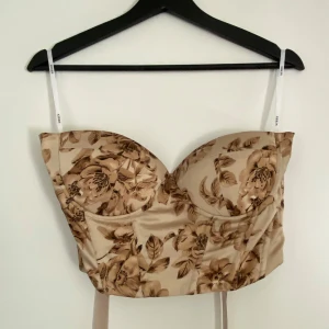 Blommig korsettopp i beige och bruna toner - Supersnygg korsettopp med blommigt mönster i beige och brunt. Toppen har smala axelband och snörning i ryggen för perfekt passform. 