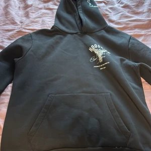 Svart hoodie från Hoodrich och svart hoodrich mjukis byxor - Svart hoodie från Hoodrich med tryck på bröstet och stor ängelgrafik på ryggen. Texten 'From Nothing To Something' och 'Product of the Streets' finns tryckt. Klassisk känguruficka och huva. Perfekt för streetwear-stil.